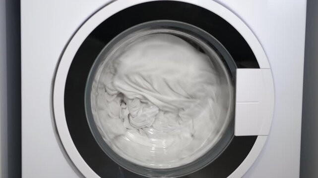 Washing machine drum spinning with clothes. Closeup of a front-loading washing machine with the drum spinning, washing white laundry and linens during a cleaning cycle. Modern household appliance