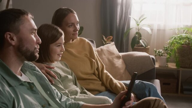 Medium side shot of happy young Caucasian family sitting together on couch at home, hugging, watching fun comedy movie, mother and father explaining plot to laughing daughter