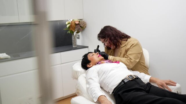 Aesthetician in a modern beauty salon carefully applying permanent makeup pigment to a woman's eyebrows with a special tool while the client lies on a cosmetology chair during a treatment