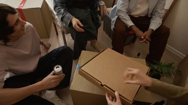 Tracking medium shot of group of four ethnically diverse friends sharing takeaway pizza among cardboard boxes in bright apartment during moving day
