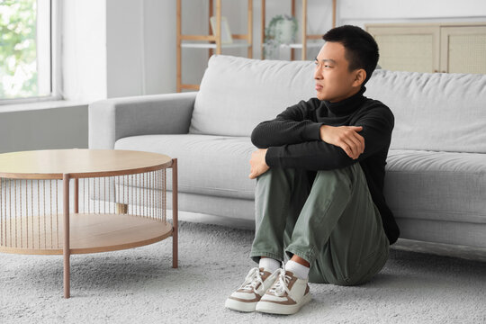 Sad young Asian man sitting on carpet at home