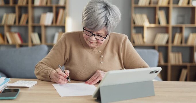 Senior woman reviews bill and plans budget. At a home desk with a tablet, she organizes finance, tracks invoice details, payment dates, saving goals and tax. Clear home budget concept today.