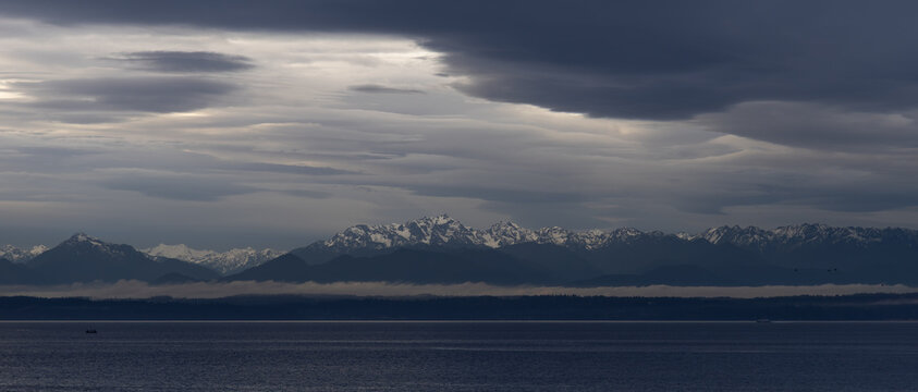 The snow capped Olympic Mountain range looms impressively across Puget Sound with moody, stormy clouds overhead. 