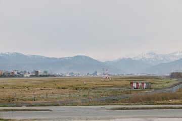 Fototapeta premium Empty rural landscape with overcast sky and distant mountains, featuring grassy fields and small buildings near a paved road. Concept of countryside, nature, and calm environment.