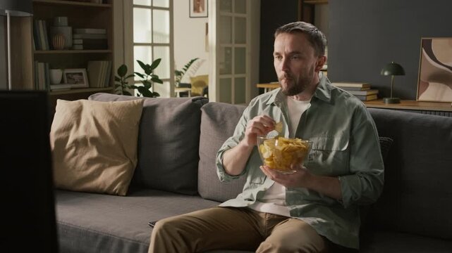 Medium full shot of young Caucasian man sitting on couch, snacking on bowl of chips, watching thriller movie on TV, staring at action on screen with shock, disbelief, then reacting with bulging eyes