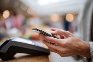 Fototapeta premium Close-up of hands holding a smartphone. Person prepares for contactless payment. Payment terminal sits on a wooden surface. Soft bokeh lights blur the background