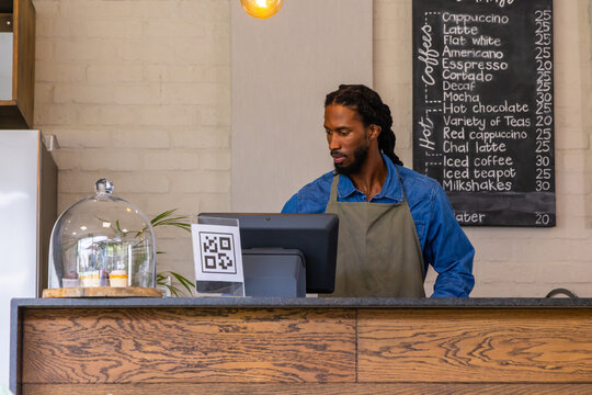 African American male barista in denim shirt apron standing behind cafe counter using POS monitor