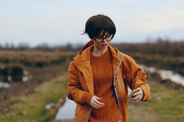 Fototapeta premium Young woman lifestyle outdoors wearing orange jacket and sweater walks on rural path with puddles. Autumn season, casual fashion and nature concept with overcast sky background.