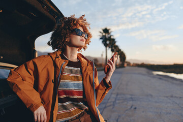 Fototapeta premium Young woman with curly hair wearing sunglasses and rainbow sweater leans on open car trunk holds smartphone outdoors on sunny day with palm trees and blue sky in background.