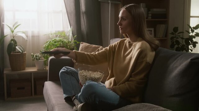 Full side of relaxed young Caucasian woman sitting down with legs on couch, choosing TV channel on streaming menu with remote, finding favorite sitcom and smiling while eating popcorn