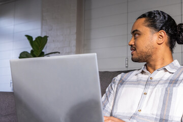Naklejka premium Asian man sitting on gray sofa in living room, typing on silver laptop, copy space