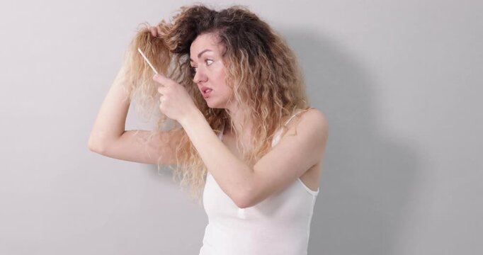 Unhappy woman trying to brush her tangled hair on light grey background