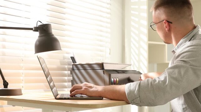 Man using printer while working on laptop at wooden desk indoors