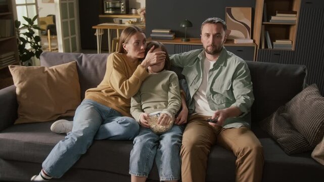 Full shot of young Caucasian family sitting together on couch, watching movie on TV, suddenly getting shocked by mature content, mother covering daughter eyes, father grabbing remote control