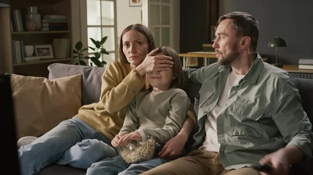 Medium shot of confused Caucasian parents and young daughter seeing inappropriate, mature or violent scene in movie, covering eyes, while caring for child mental health