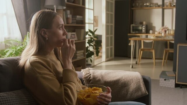 Medium panning side shot of young woman setting down on couch after doing domestic chores, snacking on bowl of chips, while watching love drama on TV with attention, focusing on storyline