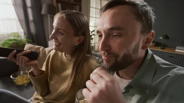 Medium close-up of Caucasian husband and wife watching hilarious sitcom show on TV, laughing aloud at gags, discussing characters and plot, while eating popcorn