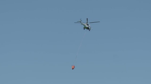 A civil protection helicopter flies across a pale blue sky carrying a suspended orange bambi bucket on a long sling line during an active aerial wildfire suppression operation with a plane in backgrou