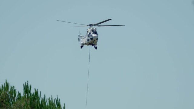 A white civil protection Super Puma helicopter flies above green treetops with a long empty sling line dangling beneath against a hazy pale blue sky during an active wildfire suppression mission