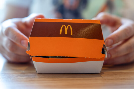 Belgrade, Serbia: July 3, 2024: Hands hold an orange McDonald's cardboard burger box on a wooden table, highlighting fast food packaging, logo branding, folded lid, and a casual indoor meal moment