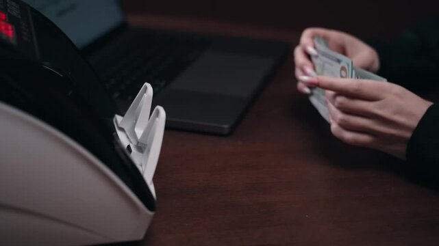 Female hand counts cash while using a money counting machine beside a laptop on a wooden table, showcasing the process of managing finances and transactions