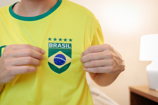 Close-up de torcedor brasileiro segurando e apontando o escudo da camisa da sele&ccedil;&atilde;o. Camisa amarela em destaque, representando orgulho, identidade e apoio em competi&ccedil;&atilde;o internacional de futebol.