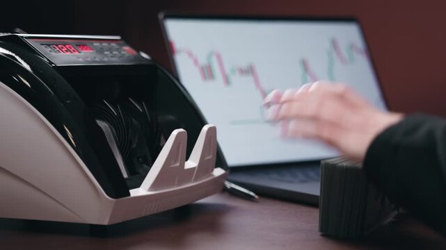 Cash counting machine processes banknotes while a person monitors financial data on a laptop screen, showcasing the connection between cash handling and financial analysis