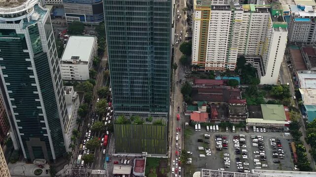 Aerial drone captures a close-up view of a blue-green glass skyscraper facade in Makati CBD with a narrow rooftop garden strip and crowded parking lot visible at the tower base in Manila.