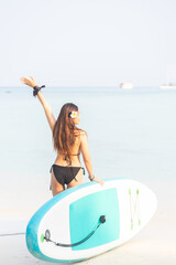 Obraz premium Young woman carrying her paddle board and paddle, walking on the beach towards the sea on a sunny day