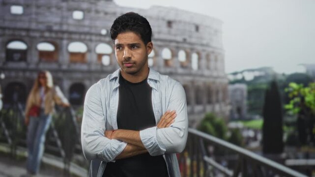 Man with arms crossed showing forearms standing by a railing at the colosseum building; quiet contemplation.