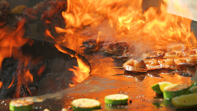 Close-up of shrimp and vegetables cooking on a hot plancha grill with large, dramatic flames. The warm, fiery lighting creates an energetic mood, perfect for food festival or barbecue concepts
