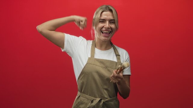 Young blonde woman makeup artist holding powder brushes flexes right arm muscle and smiles against red studio wall; confidence empowerment celebration.