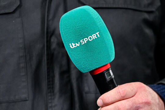 Hand holding a sports microphone with green ITV Sport cover, close-up shot of broadcasting equipment during a media interview.