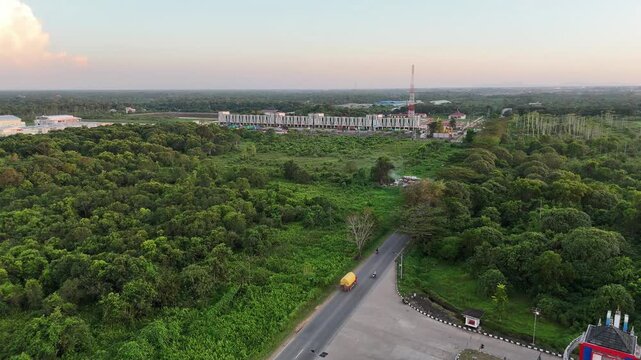 A cinematic aerial shot pans across the main road and lush forest toward a striking gate, a modern commercial complex, and a telecommunications tower at dusk.
