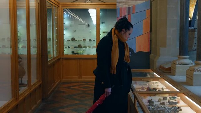 A woman examining mineral specimens inside a museum display case, holding a closed red umbrella while standing beside illuminated exhibits in a historic natural history gallery
