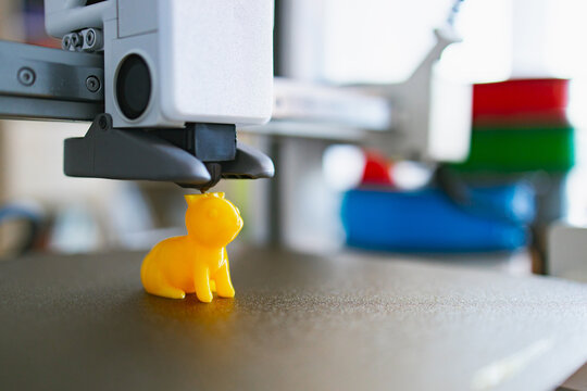 A 3D printer nozzle deposits molten plastic to create a small, bright yellow rabbit figurine on its build plate