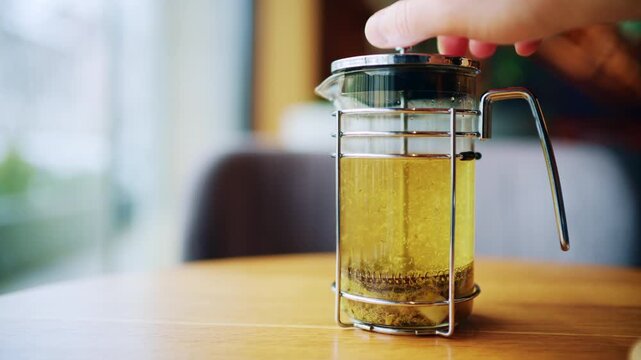 Woman pressing French press to brew herbal tea in glass teapot on wooden table in cozy interior