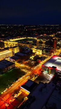 A vertical drone shot captures the vibrant glow of Joliet, Illinois, city streetlights and modern office buildings against the dark night sky during winter.