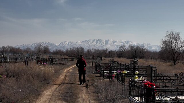 a man walks into the distance along a dirt road through an old rural cemetery against the background of graves and mountains in the snow, close-up