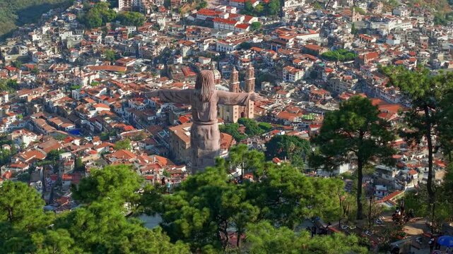 Aerial orbit establishing shot from the Monumental Christ of Taxco overlooking the town of Taxco with the Santa Prisca temple and colonial architecture of white houses with tiled roofs, Mexico