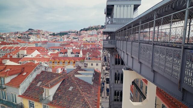 The ornate iron structure of the Santa Justa elevator frames a sweeping panorama of Lisbon, Portugal, with terracotta rooftops cascading toward the hilltop Castelo de Sao Jorge under a cloudy winter s