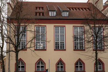 Obraz premium Beige building facade with red window frames and a large leafless tree in front on a quiet winter day.
