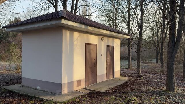 A functional small building with brown doors sits on the edge of a quiet, frost-covered forest. The long morning shadows and crisp air create a feeling of lonely utility in the winter park.