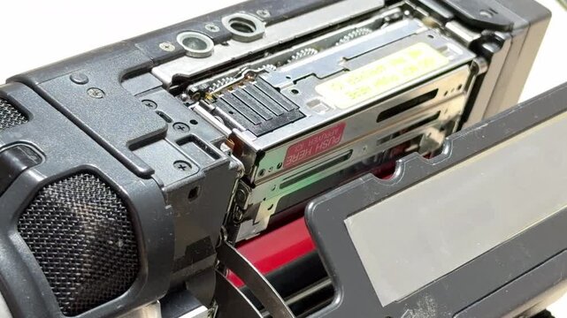 A vintage digital camcorder opens its side compartment to eject a MiniDV tape. The motorized mechanism produces a high-pitched mechanical whir followed by a metallic clicking sound