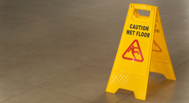 A yellow plastic folding sign with a caution wet floor warning placed on a textured grey floor mat to prevent slips and falls in a commercial indoor setting.
