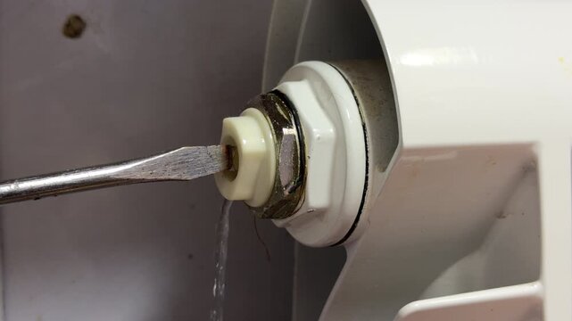A flat-head screwdriver is inserted into a plastic bleed valve on a white radiator. Water is spraying out in a thin stream, indicating the radiator is being bled.