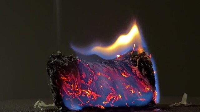 A small bundle of fiber smolders with deep red embers and a vivid blue flame. The glowing heat creates a sense of raw energy and quiet intensity. It is a mesmerizing and powerful sight.
