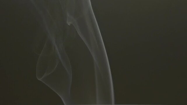delicate stream of white smoke drifts upward, creating soft wispy patterns. The vapor disperses into a solid dark grey environment, suggesting a recently extinguished flame or heat source.
