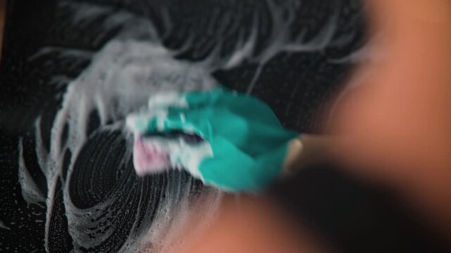 A hand with a green glove scrubs a surface using a soapy sponge. The action demonstrates proper cleaning techniques and personal safety measures in a cleaning environment