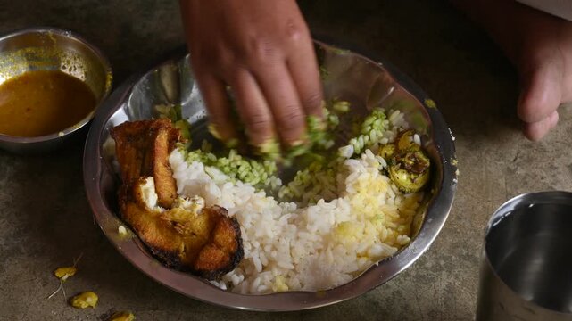 A person is eating piping hot white rice and fish curry. It is served alongside fried fish and roasted bitter gourd. Homemade, traditional Indian food. Indian meal, served in a traditional steel plate